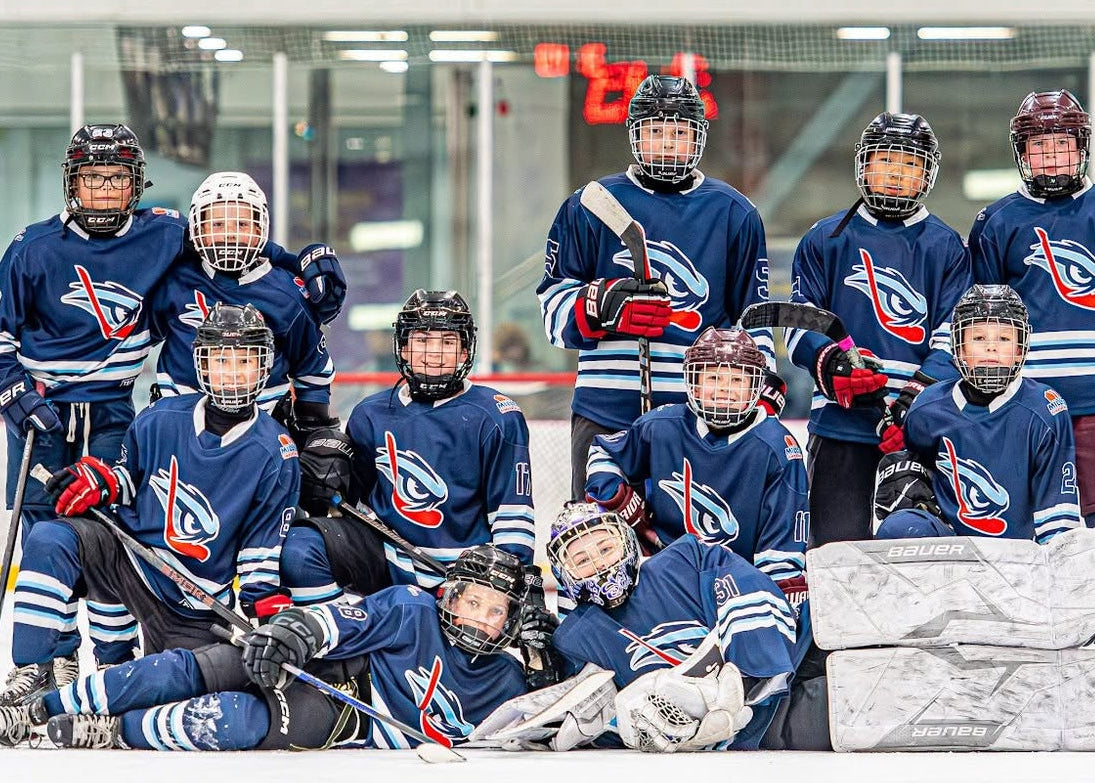 A group of youth hockey players posing on an indoor ice rink, wearing matching dark blue jerseys with white and red accents, black helmets, and holding hockey sticks.